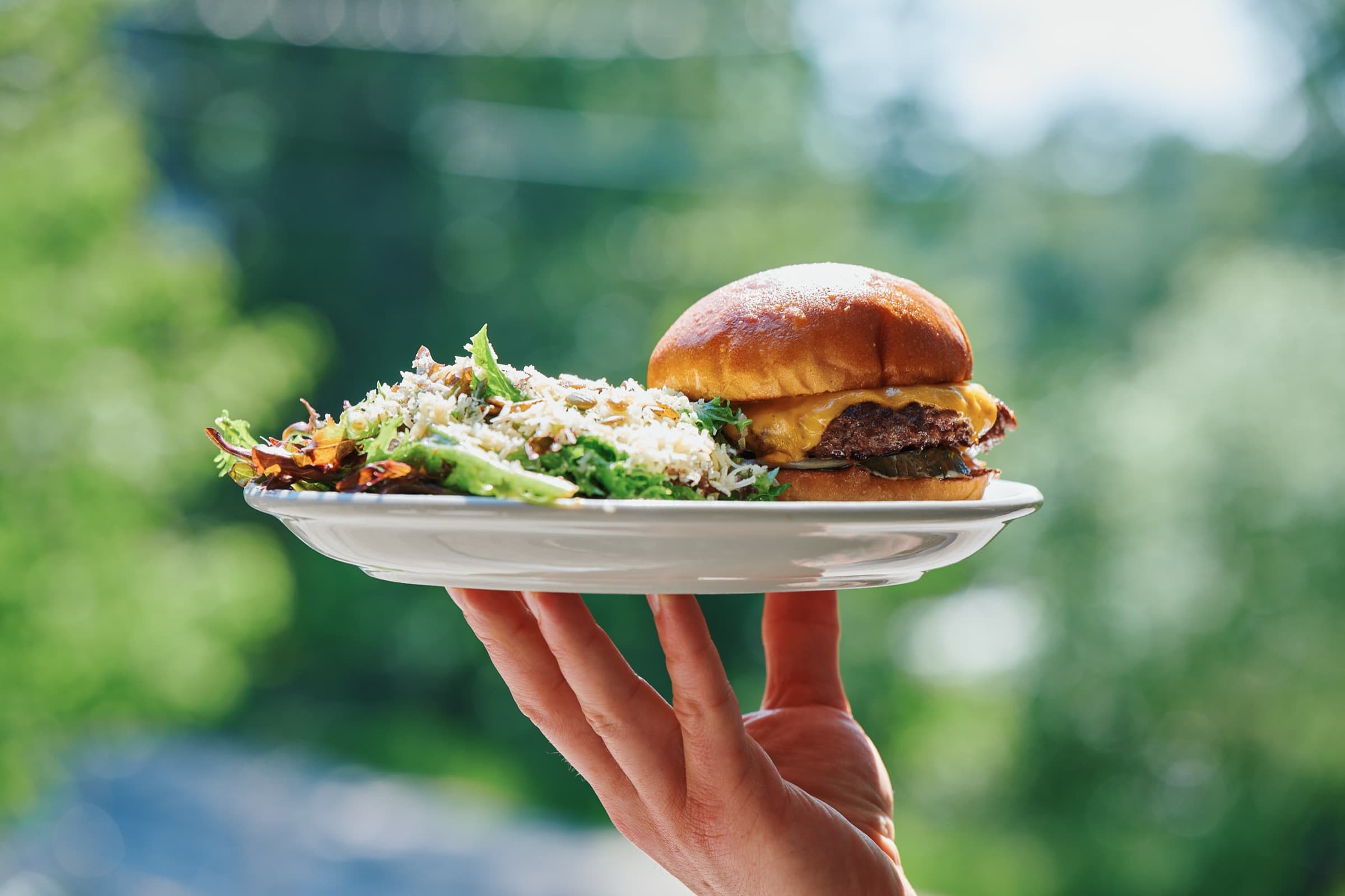 Une assiette avec cheeseburger et salade verte levée vers la lumière, la verdure des Laurentides en arrière-plan