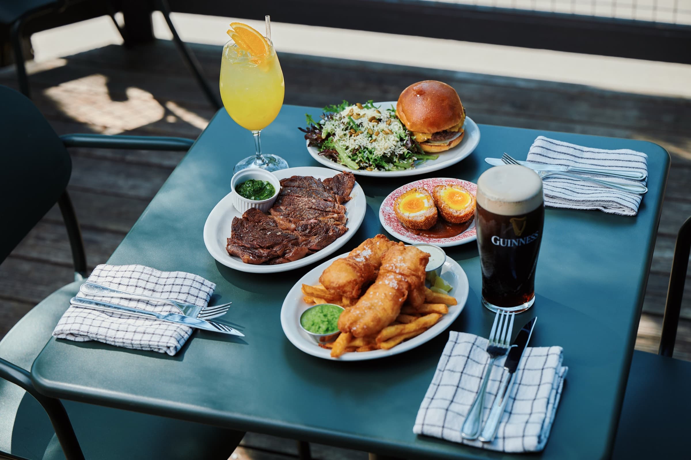 Overhead view of a terrasse table with grilled steak, soft-yolk scotch eggs, fish and chips, a burger with salad, a cocktail and a pint of Guinness.
