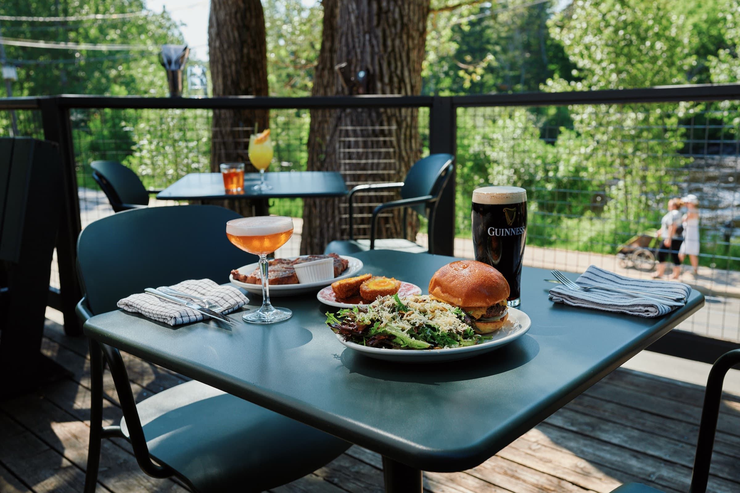 MacTaverne's terrasse in summer: a table set with a cheeseburger, a Guinness and a cocktail, with trees and the trail in the background.