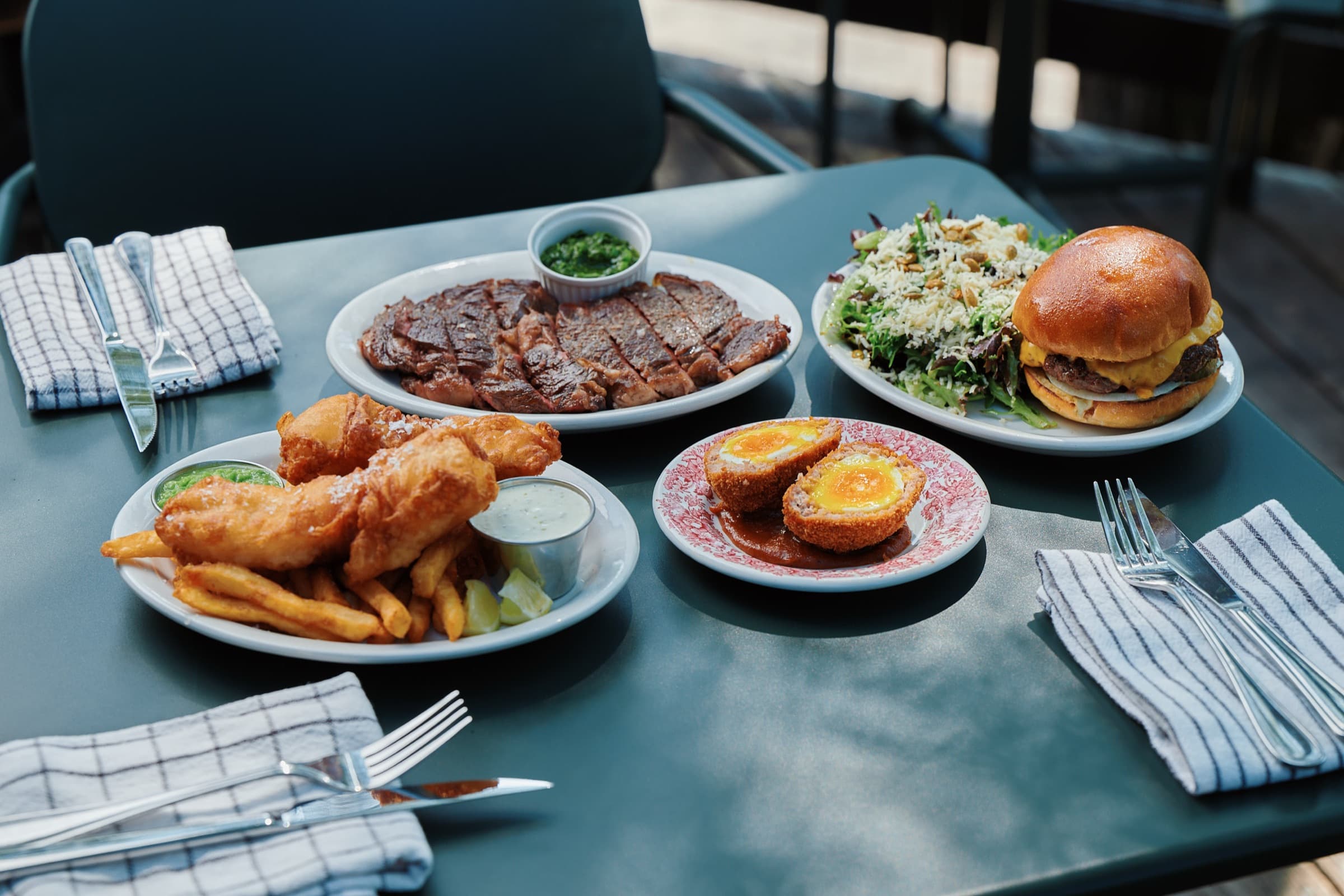 Une table ensoleillee garnie de steak grille tranche, d'un cheeseburger avec salade, d'un scotch egg et d'un fish and chips avec puree de pois.
