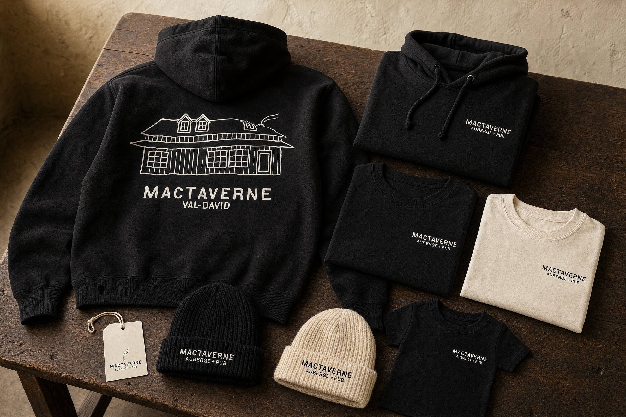 A MacTaverne hoodie, tees and tuques in black and cream, laid out on a wooden table.