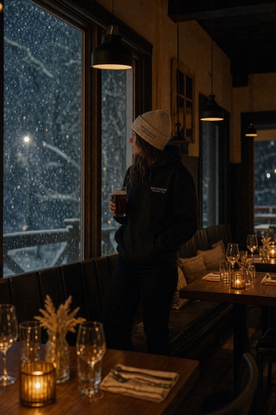 On a snowy evening, a person in a cream tuque and a black MacTaverne hoodie holds a dark beer and watches the snow fall through the big dining room window, candlelit tables set around.