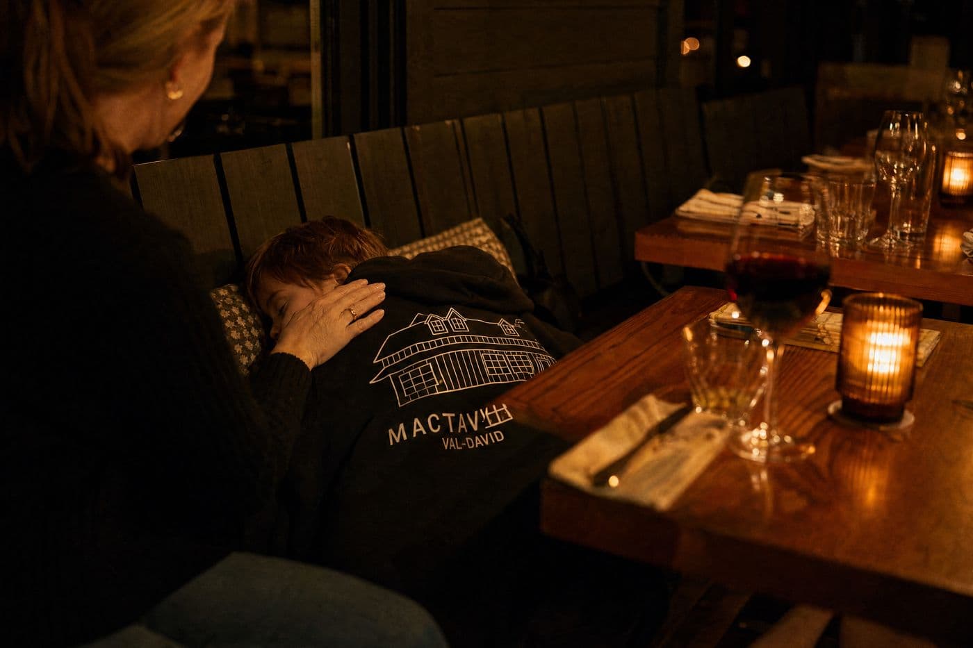 In the evening dining room, a child sleeps on the banquette under a black MacTaverne Val-David hoodie while a woman rests a hand on the child's back; candles and a glass of red wine on the table.