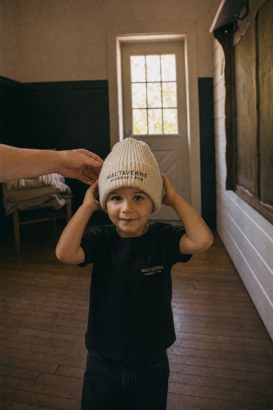 In the entrance hall, a young child in a black MacTaverne Auberge + Pub tee adjusts a ribbed cream tuque while an adult's hand helps.