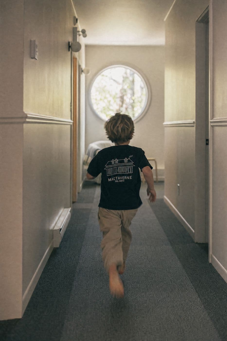 Seen from behind, a child in a black tee with the house drawing and MACTAVERNE VAL-DAVID runs down a hallway toward a round window.