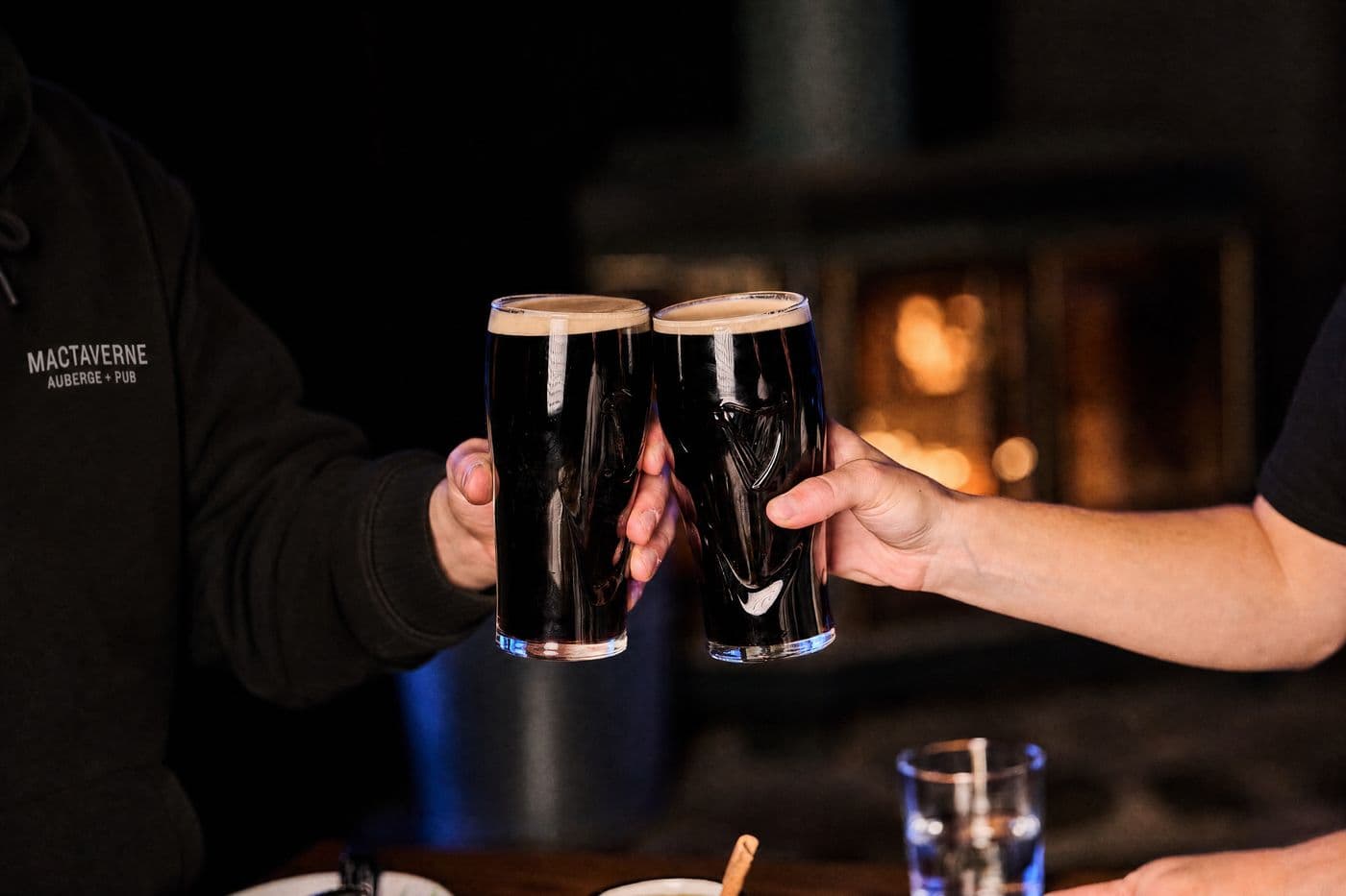 Close-up of two hands clinking pints of stout in front of the fireplace, one in the sleeve of a black MacTaverne Auberge + Pub hoodie and the other in a black tee sleeve.