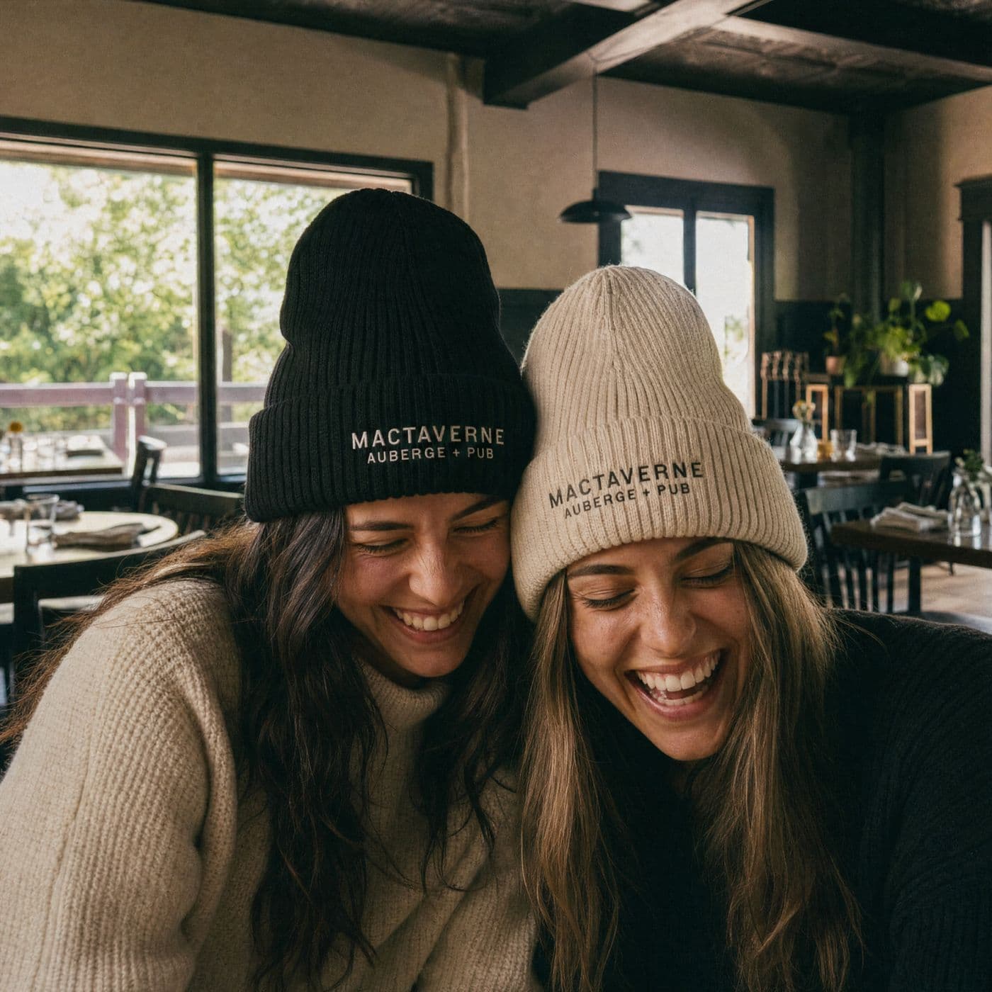 Two women in black and cream MacTaverne Auberge + Pub tuques laugh with their heads together in the sunlit dining room.