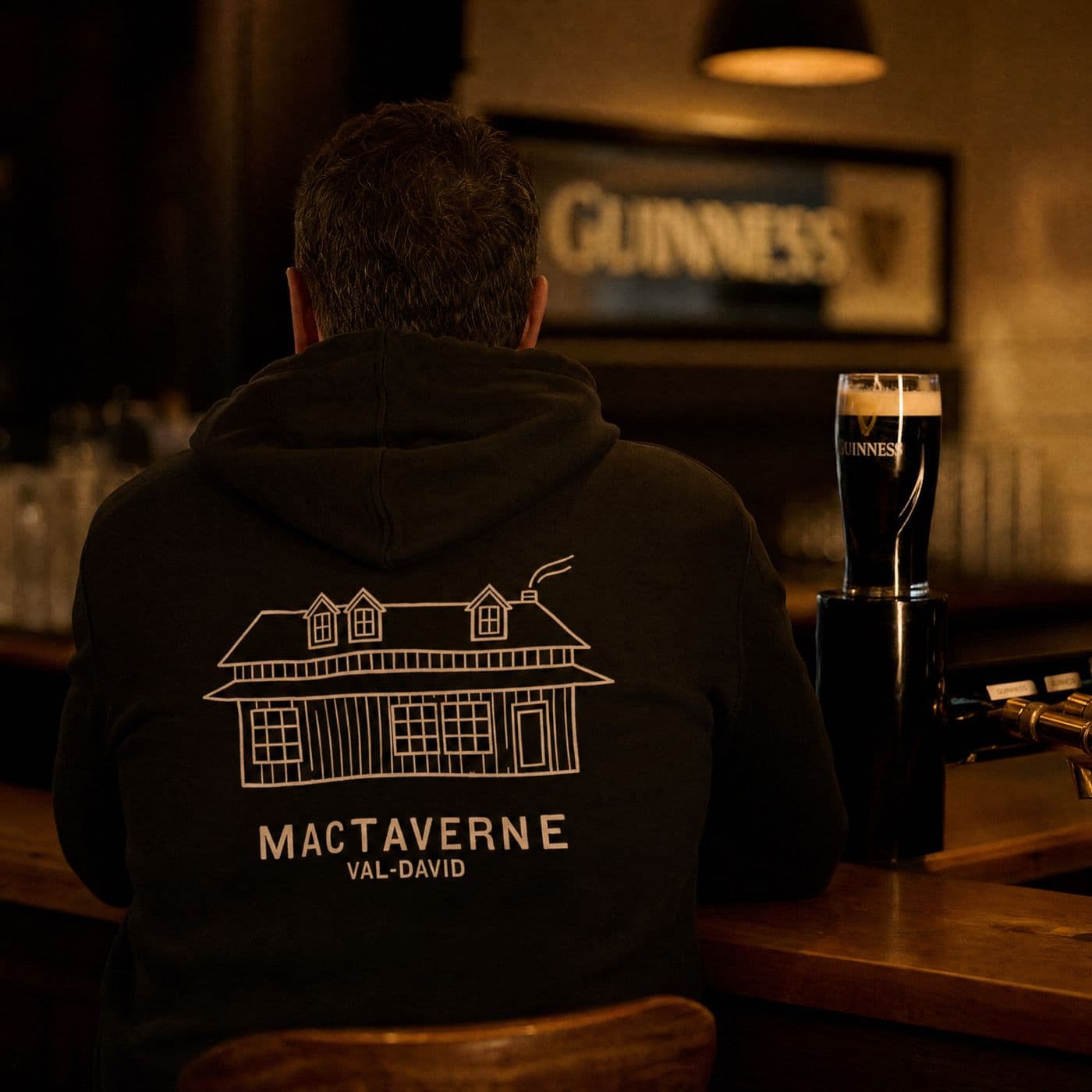 Seen from behind at the bar, a man wears the black hoodie printed with the house drawing and MACTAVERNE VAL-DAVID; a pint of stout waits beside him.
