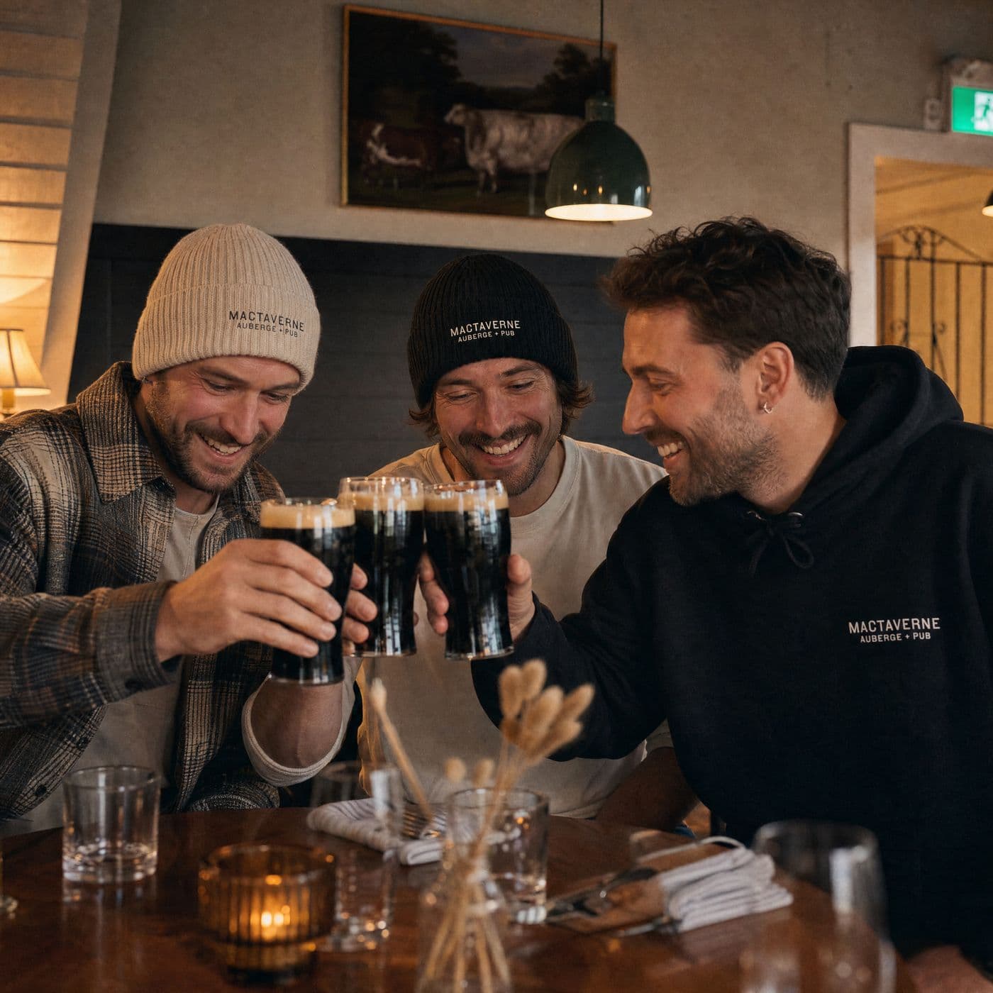 Three people clink pints of stout: a cream tuque with a plaid shirt, a black tuque with a cream tee, and a black MacTaverne Auberge + Pub hoodie.