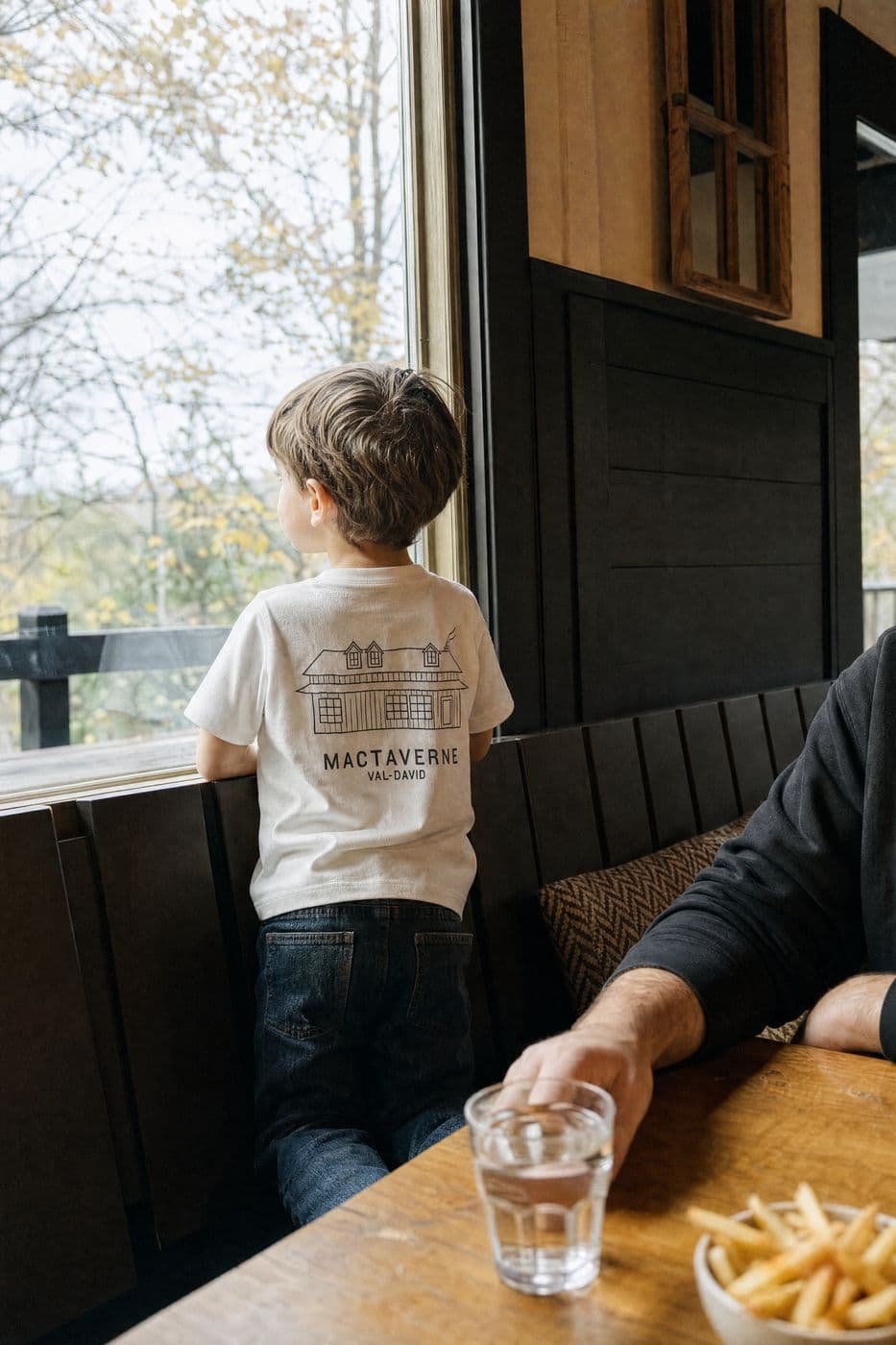 Seen from behind, a young boy in a cream MacTaverne Val-David kids tee looks out the dining room window, beside a glass of water and a bowl of fries.