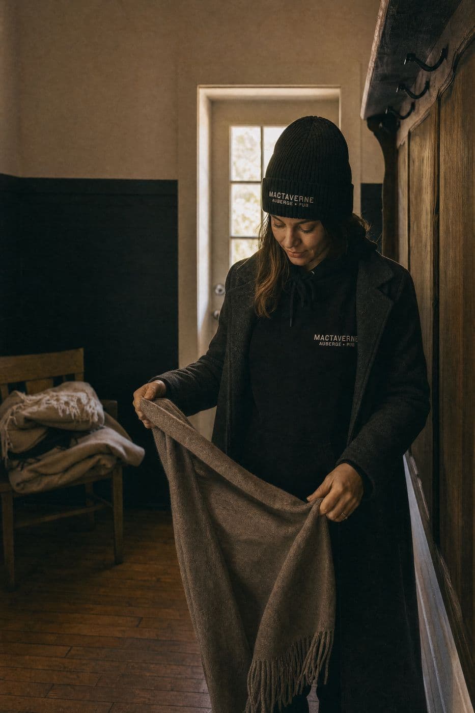 In the entrance hall, a woman in a black tuque, a black MacTaverne Auberge + Pub hoodie and a long grey coat unfolds a brown wool blanket.