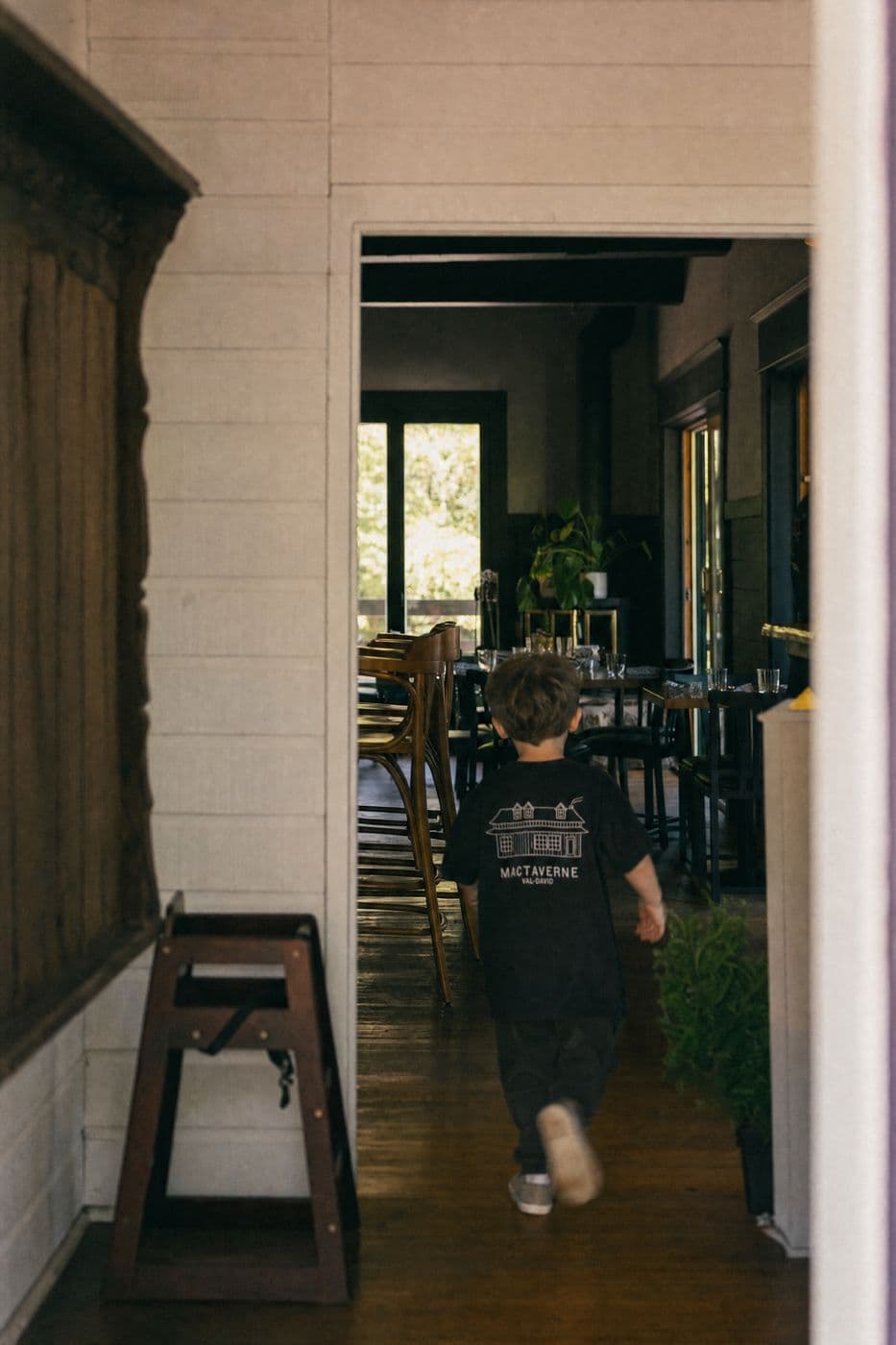 Seen from behind, a young child in a black tee with the house drawing and MACTAVERNE VAL-DAVID walks from the entrance toward the dining room.
