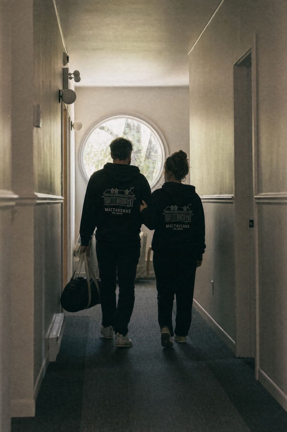 Seen from behind in a hallway with a round window, a man with a duffel bag and a woman walk arm in arm in black hoodies printed with the house drawing and MACTAVERNE VAL-DAVID on the back.