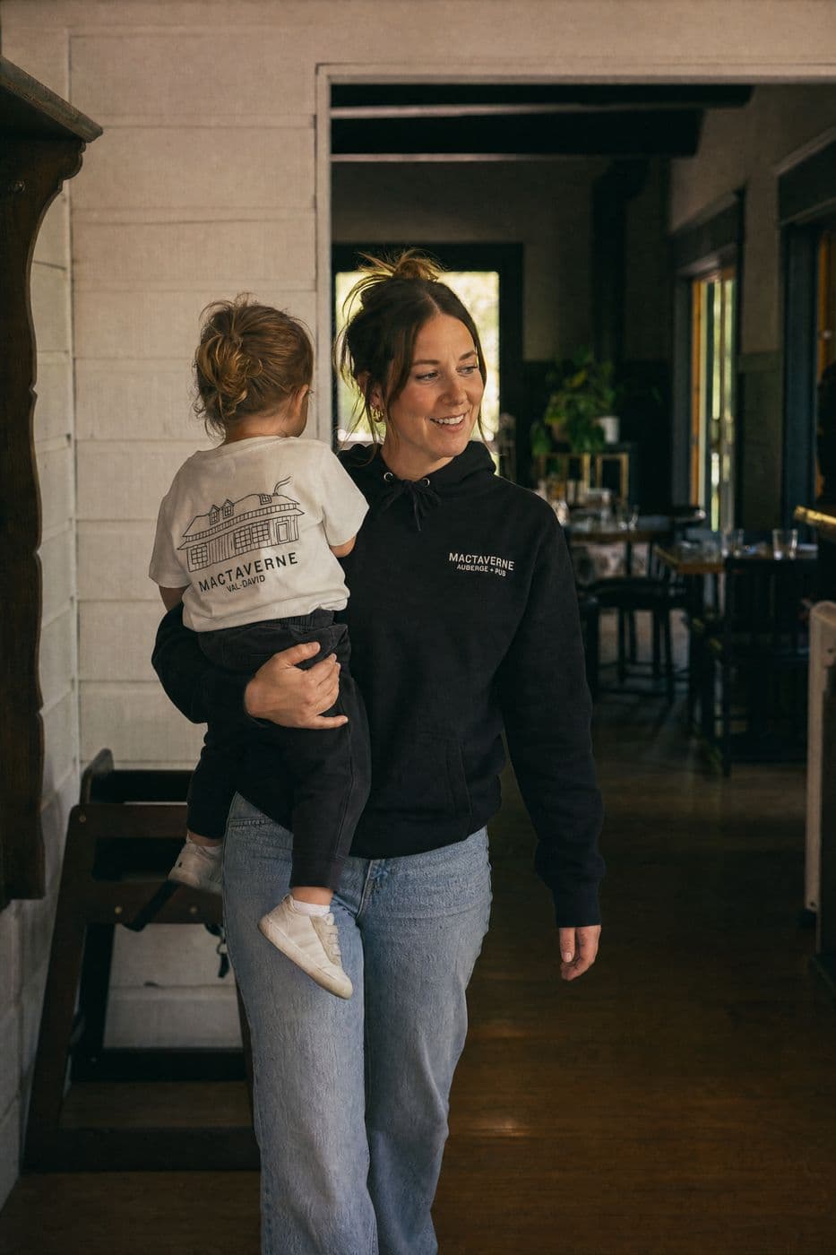 In the entrance hall, a smiling woman in a black MacTaverne Auberge + Pub hoodie carries a toddler whose cream tee shows the house and MACTAVERNE VAL-DAVID on the back.